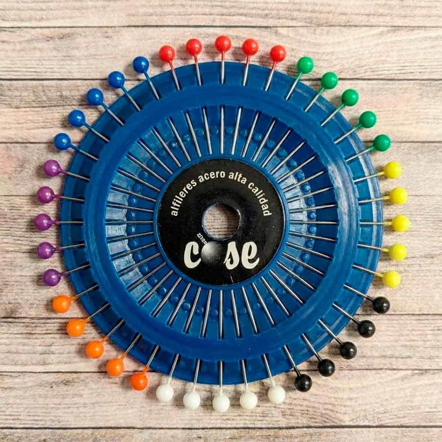 Disco azul porta-alfileres con 40 alfileres de cabeza redonda multicolor sobre fondo de madera clara.