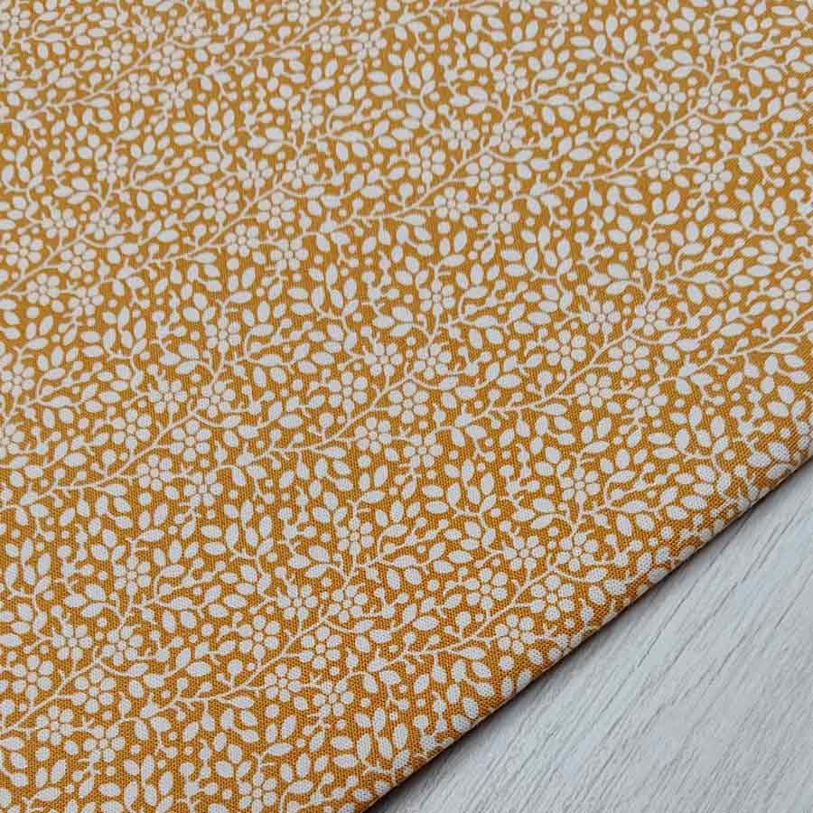 Tela de algodón americano con estampado de pequeñas flores y hojas blancas sobre fondo color mostaza, especial para patchwork.